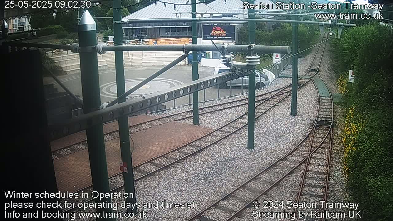 A tram station's curving tracks are seen from an elevated perspective, with a vehicle parked nearby, and lush greenery bordering the area.