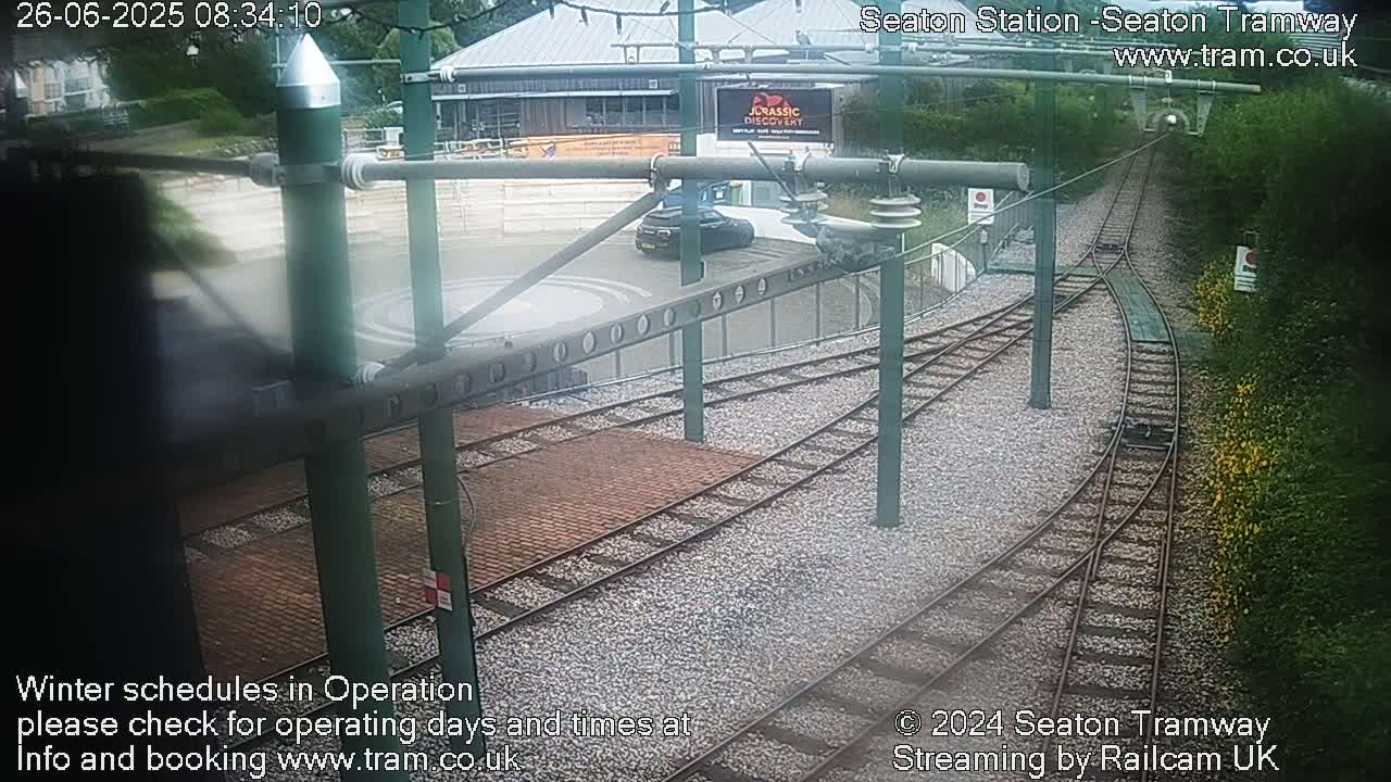 A tram station with converging train tracks, a car parked nearby, and surrounding greenery under overcast skies.