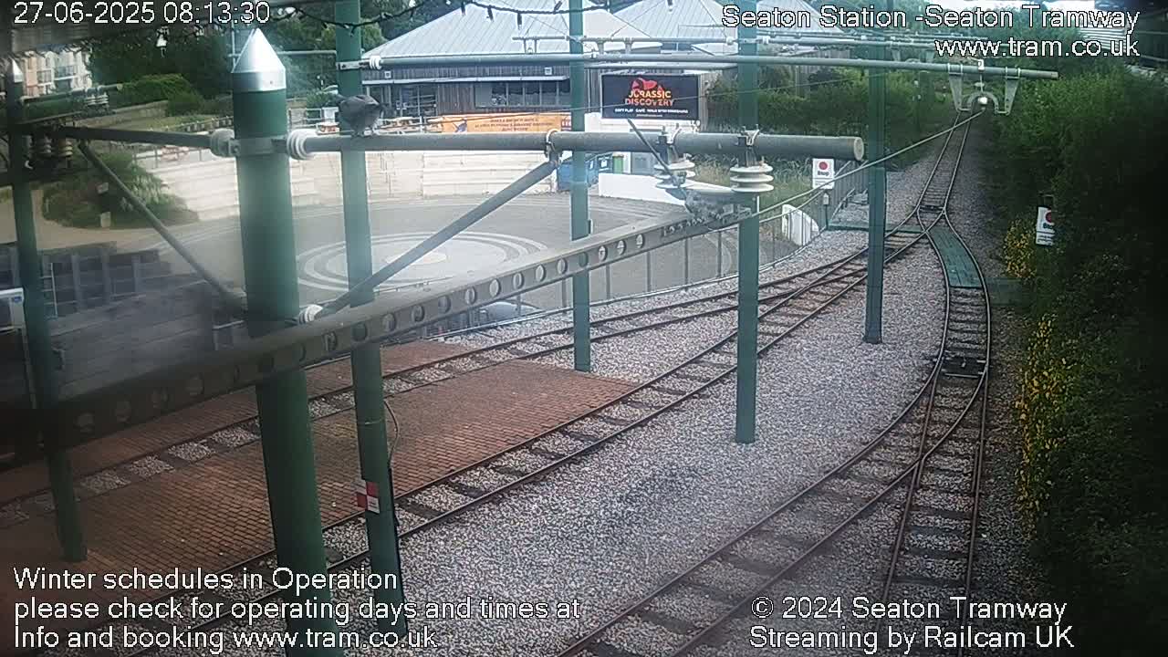 Tram tracks curve through a station area next to a building and greenery under a partly cloudy sky.