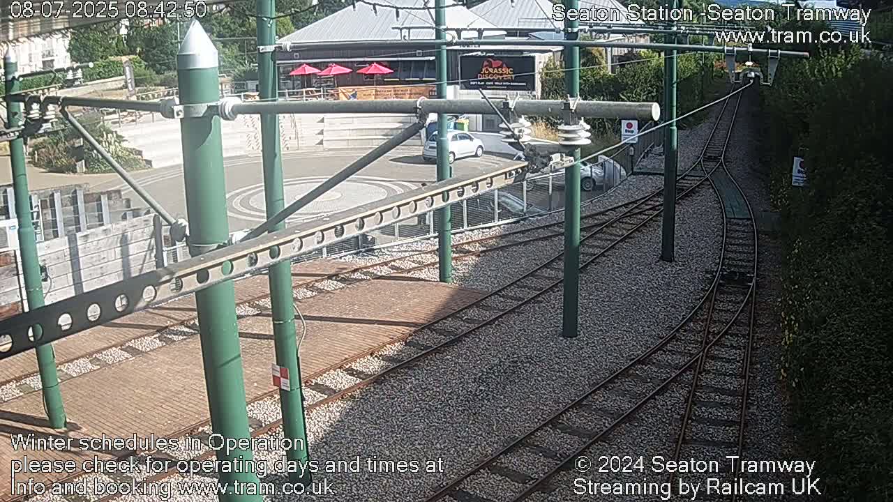 A high-angle view shows a sunny day at a tram station with train tracks branching off, a roadway, and a building in the background.