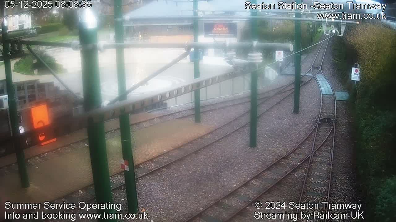 An empty tram station, featuring multiple tracks, wet platforms, and green support poles for overhead lines, is seen on a damp and overcast day.