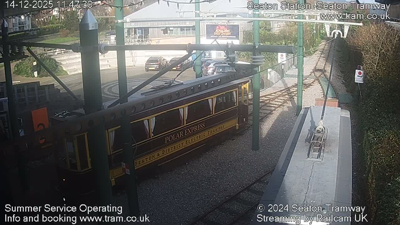 An empty tram station, featuring multiple tracks, wet platforms, and green support poles for overhead lines, is seen on a damp and overcast day.