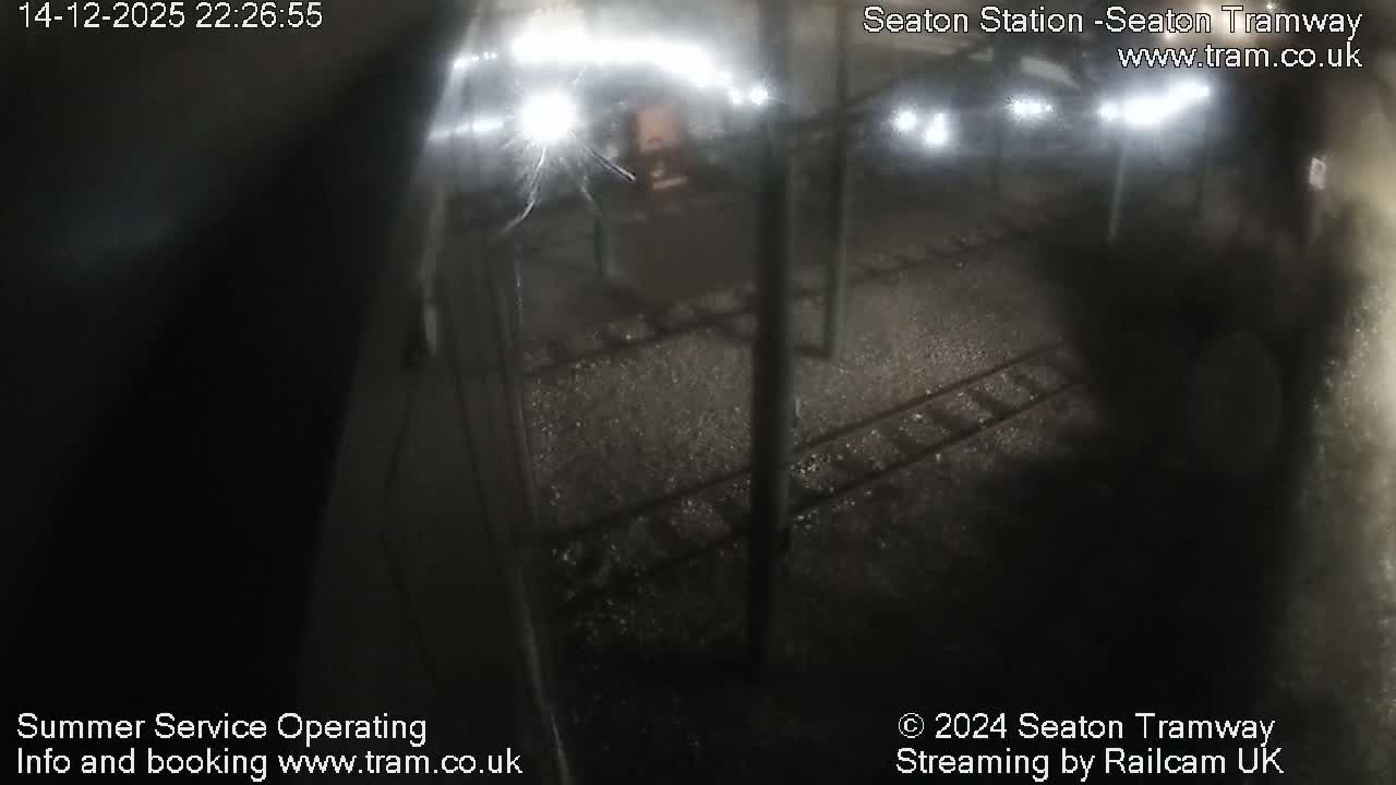 An empty tram station, featuring multiple tracks, wet platforms, and green support poles for overhead lines, is seen on a damp and overcast day.