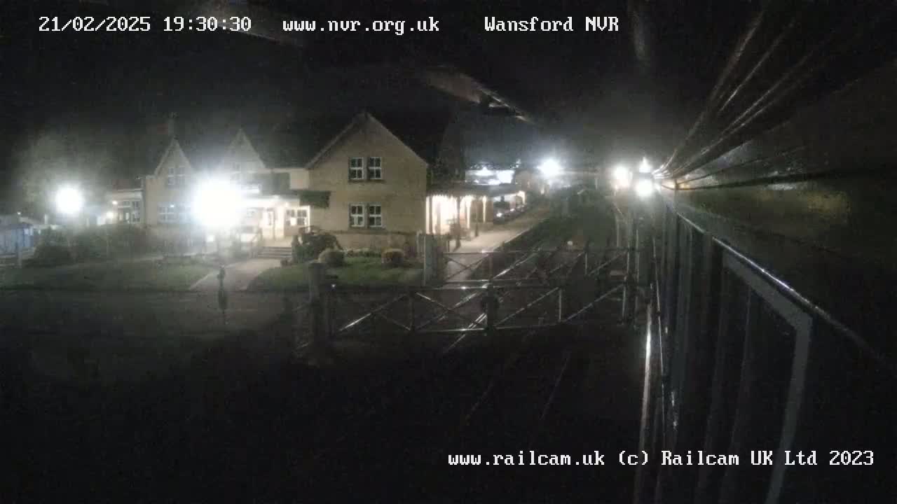 A nighttime view of a building illuminated by lights, with a dark, possibly rainy, outdoor setting and a portion of what appears to be a train car visible in the foreground.