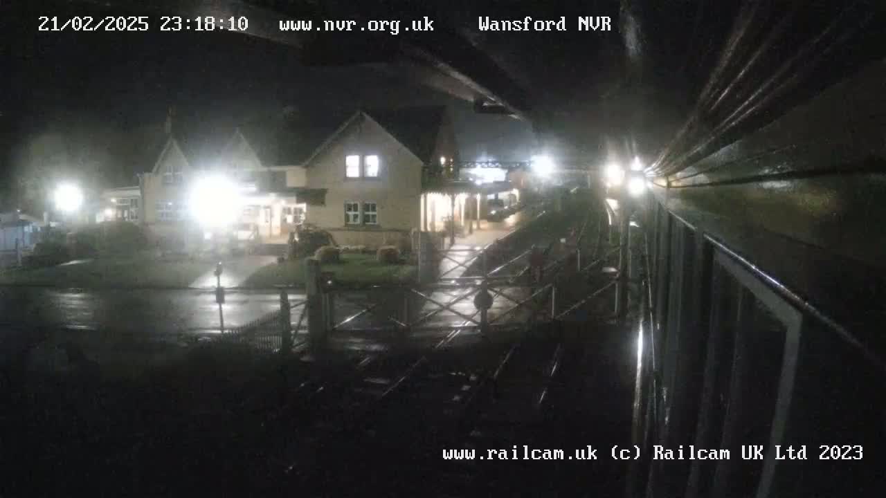 A nighttime view of a train station in the rain, showing a building and a railway crossing.