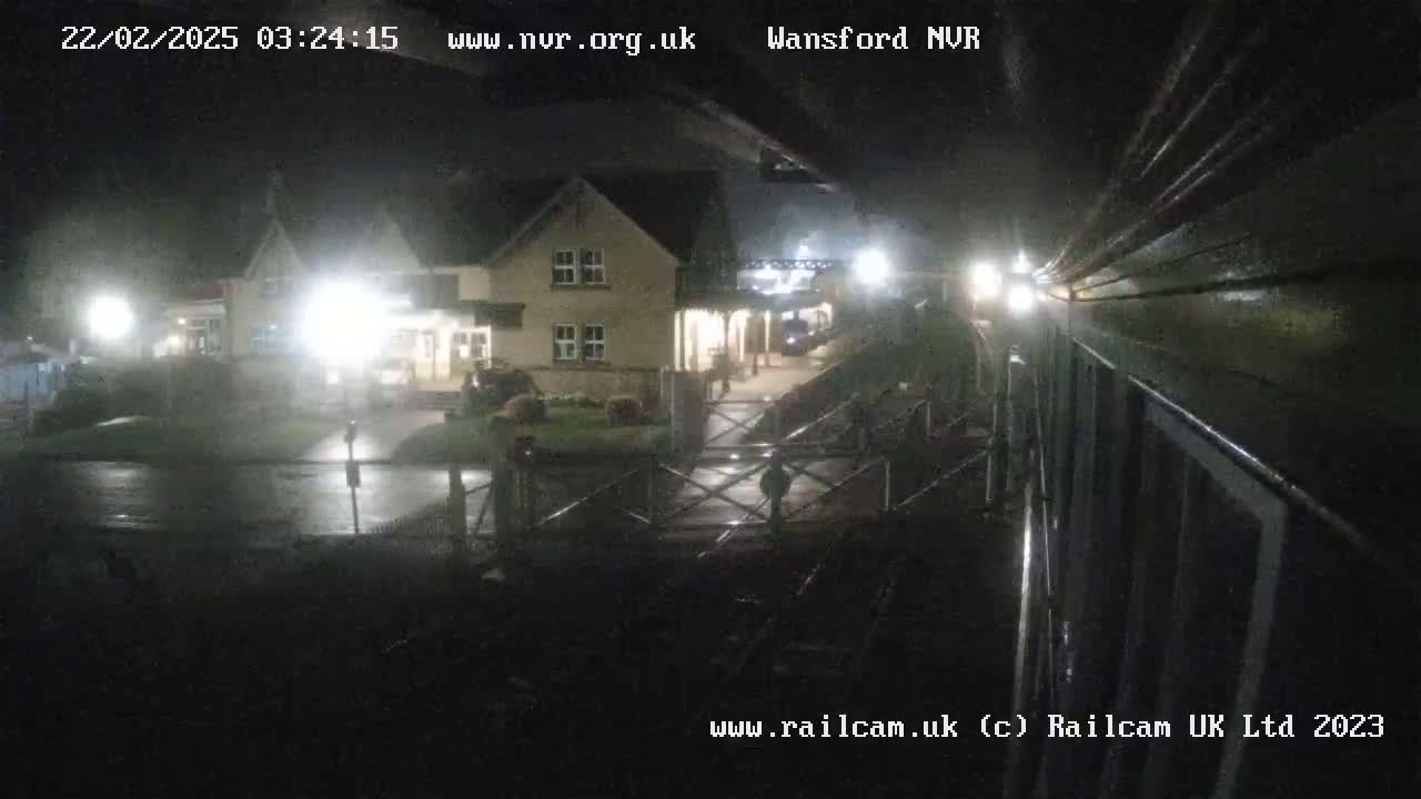 A nighttime view of a rain-slicked railway station, showing a building and a crossing gate under dim lighting.