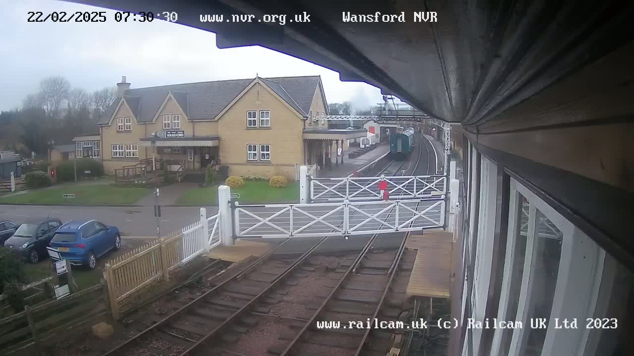 A train is departing from a station on an overcast day, passing a level crossing with cars parked nearby.