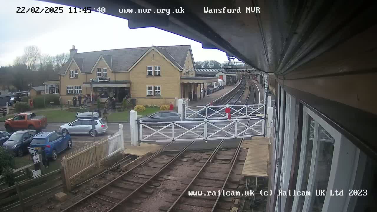 A view from a train shows a railway crossing with cars parked nearby, a station building, and overcast skies.