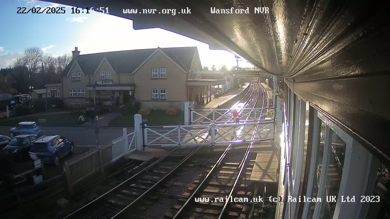 A sunny view from a train platform shows a railway crossing gate, train tracks, a station building, and several parked cars.