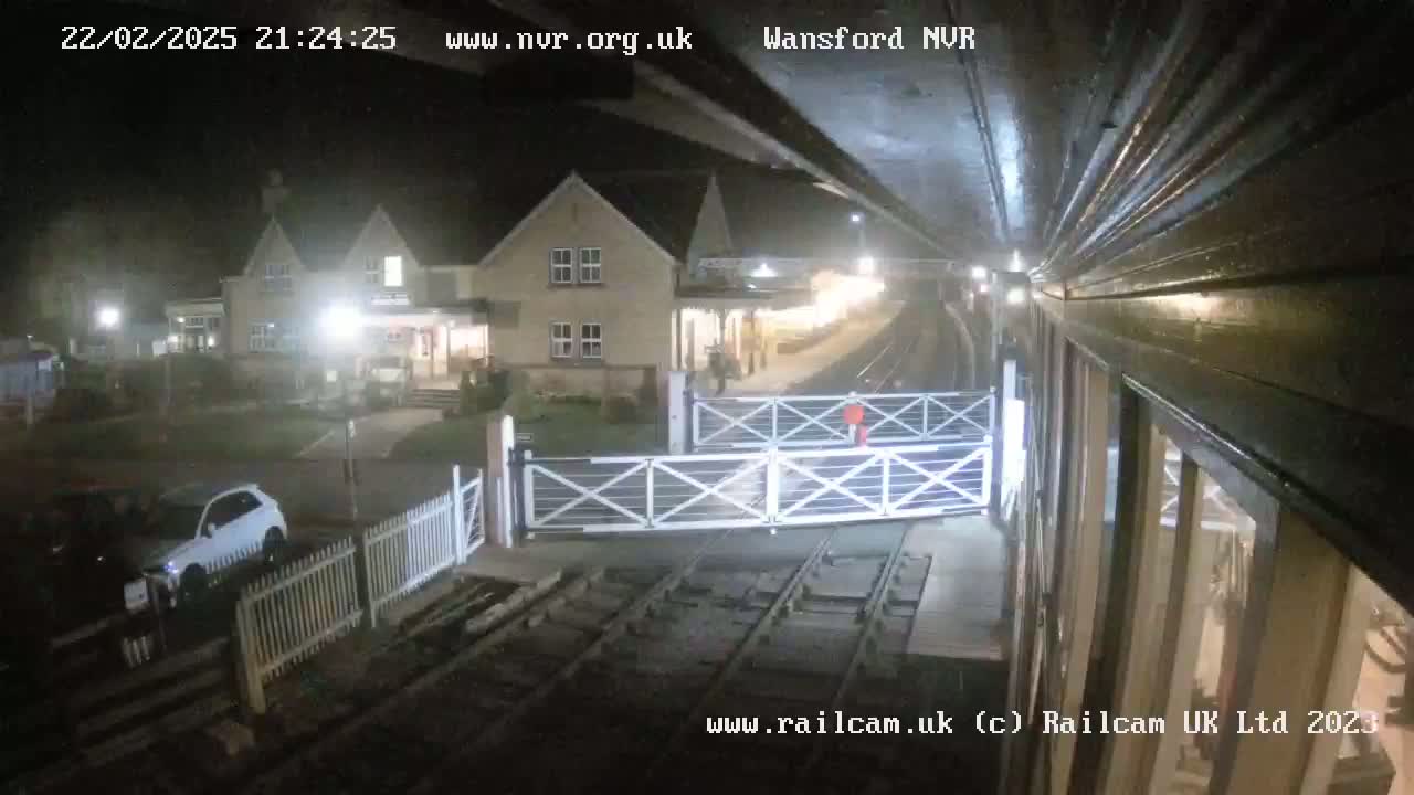 A nighttime view of a train station with closed gates, a building, and a couple of cars parked nearby under hazy conditions.