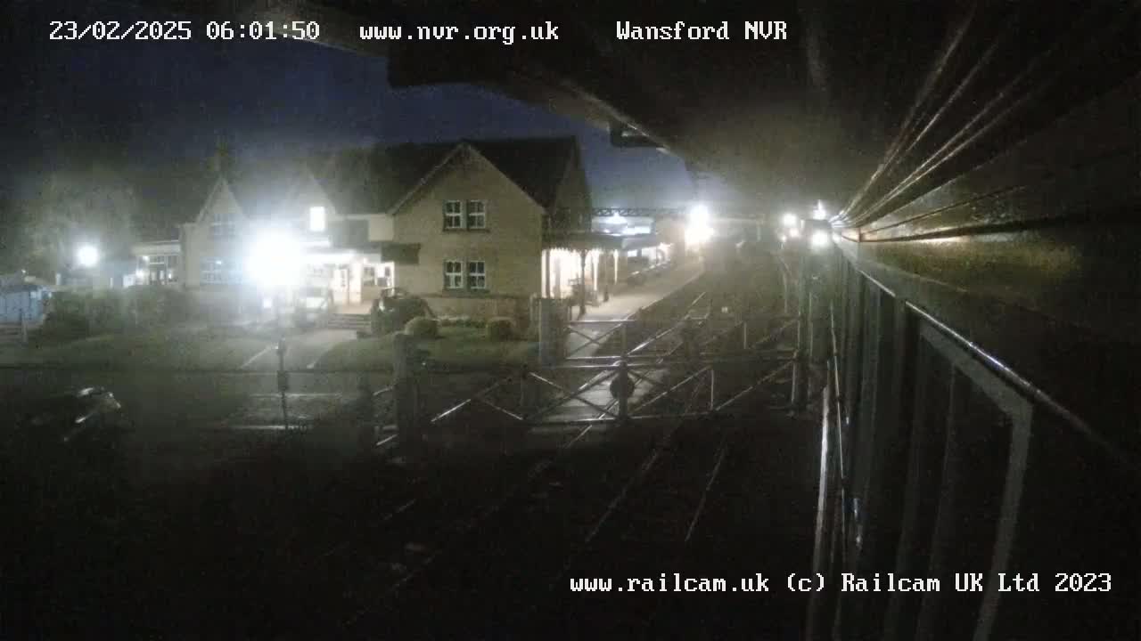 A nighttime view from a train shows a dimly lit station building and a gated crossing.