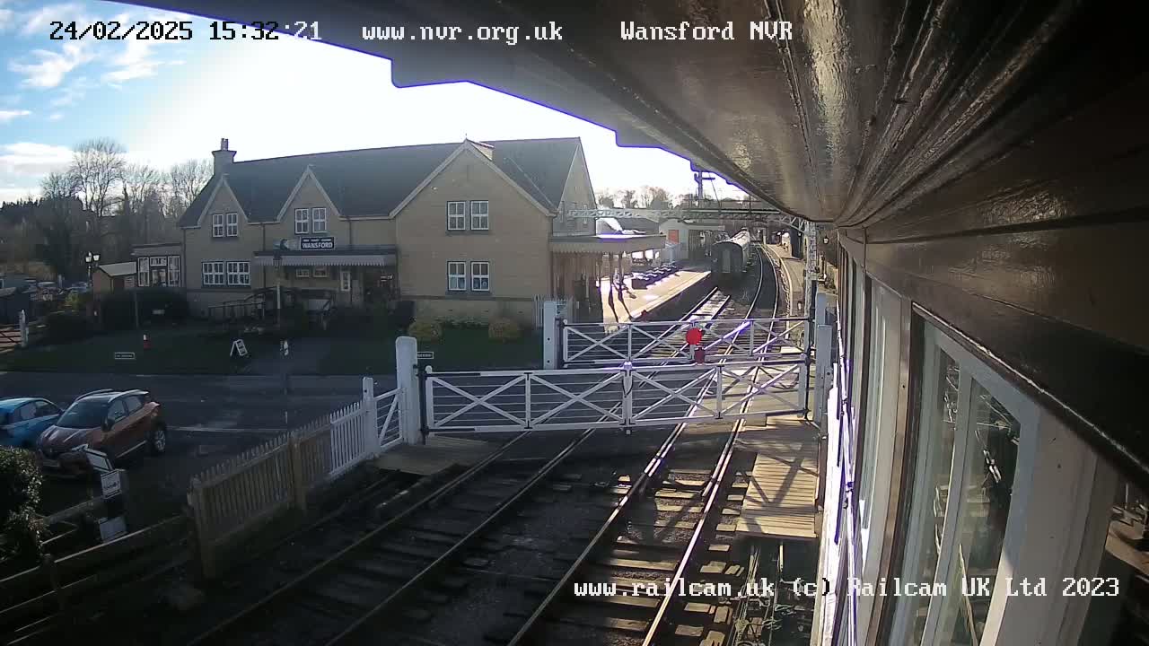 A sunny view from a train overlooking a railway station with a building, cars in a parking lot, and a person walking near the tracks.