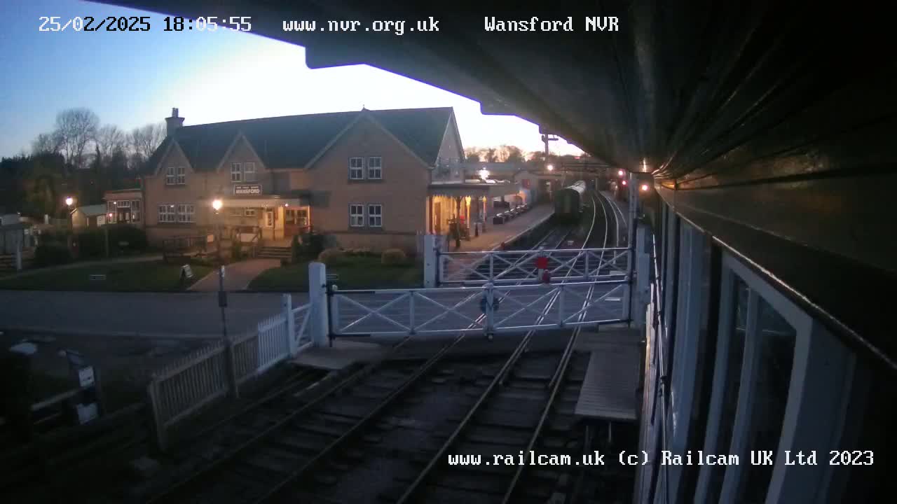 A train station at twilight, with a two-story building and a train on the tracks.