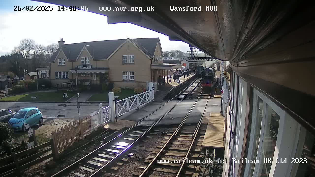 A steam train arrives at a station on a sunny day, with a crowd of people on the platform and cars parked nearby.