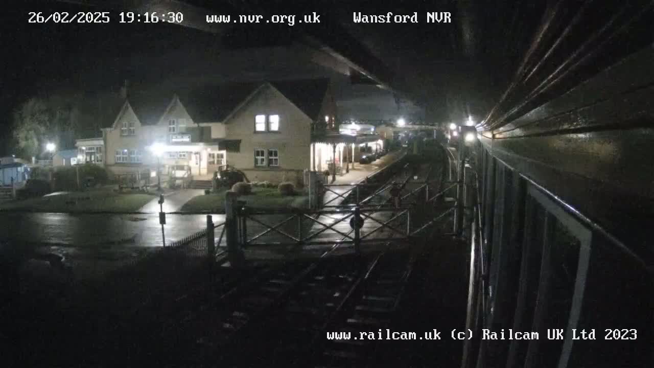 A nighttime view from a train shows a wet station platform and a building with lights on, under a dark, cloudy sky.