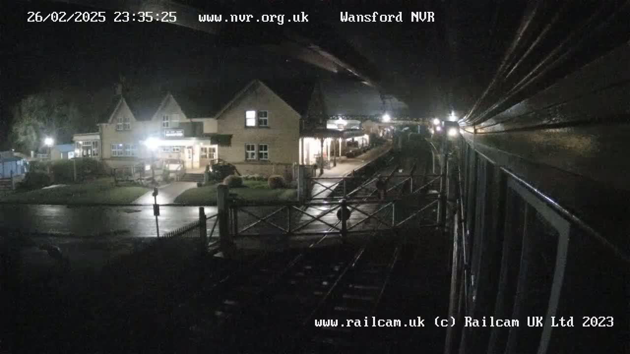 A nighttime view from a train shows a wet, dimly lit train station with a building and platform visible.