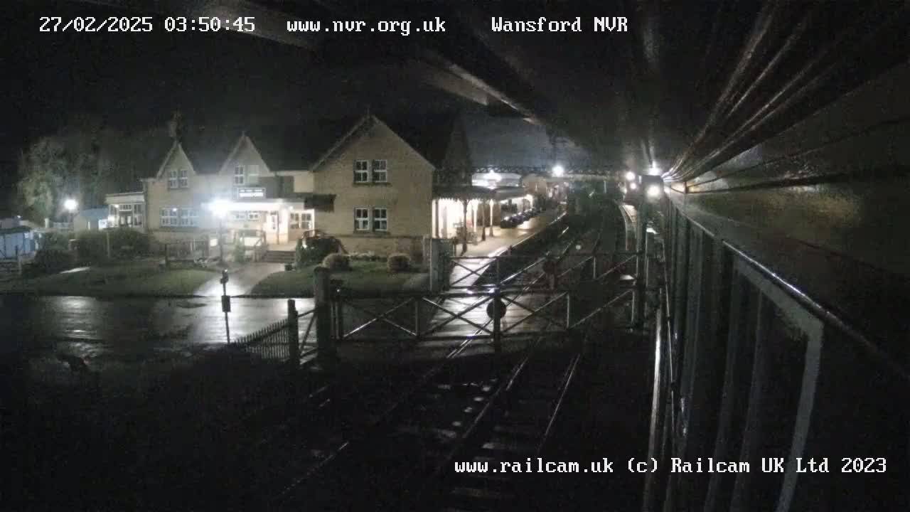 A nighttime view of a train approaching a wet station, with a building illuminated in the background.