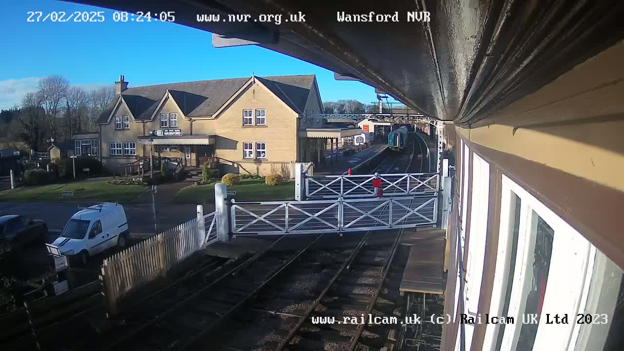 A train is visible at a railway station on a sunny day, with a two-story building and a white van parked nearby.