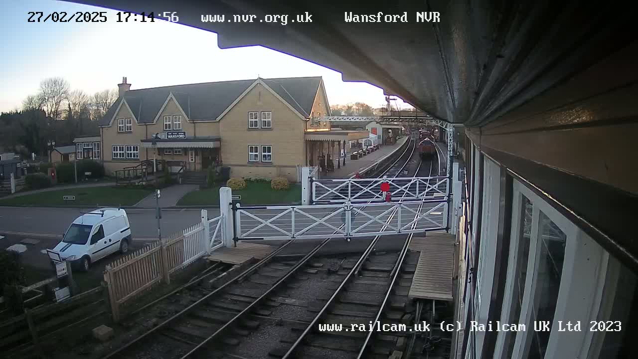 A train is arriving at a station, near a white van and a stone building, on a partly cloudy day.