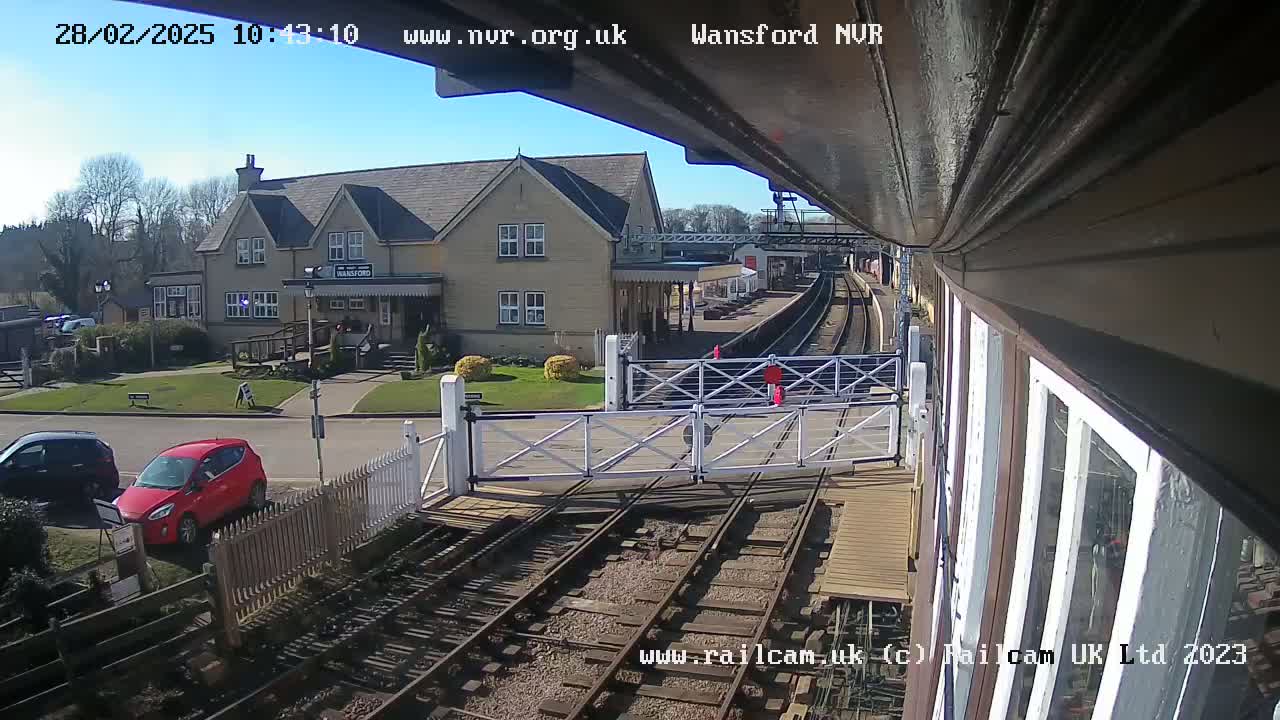 A sunny view from a train platform shows a building, a parking lot with cars, and railroad tracks with crossing gates down.