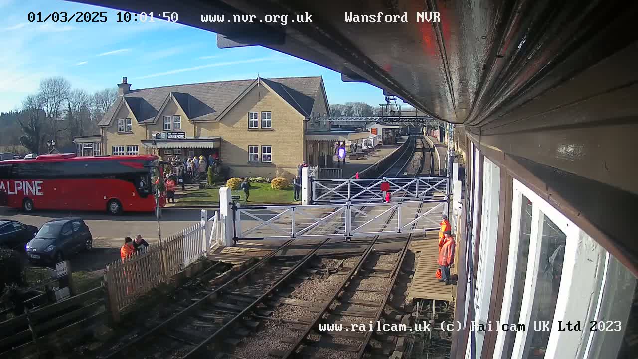 A sunny day at a train station shows a red bus, several people, a level crossing, and train tracks.
