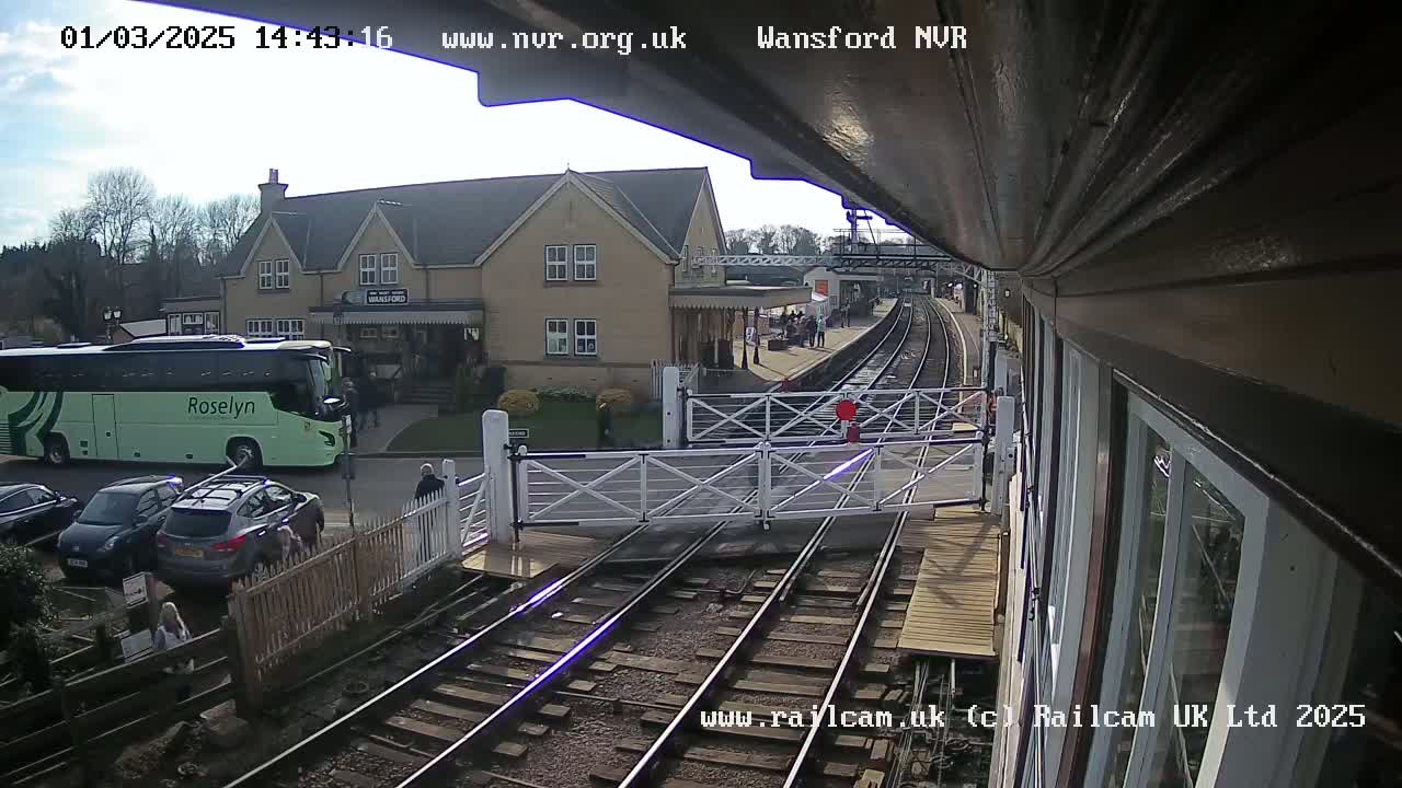 A sunny view from a train shows a railway station with a light green bus, several cars, and people on a platform next to a building.