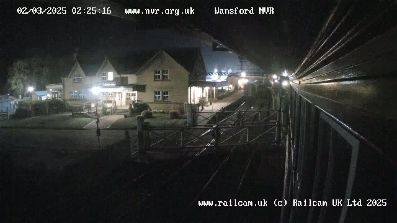 A nighttime view from a train shows a two-story building with lights on, next to a train station platform and tracks.
