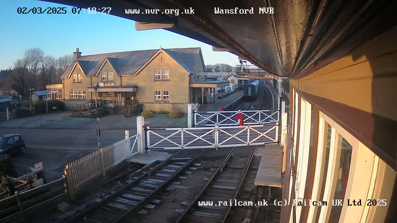 A train is at a station on a sunny morning, with a building and crossing gates visible in the foreground.