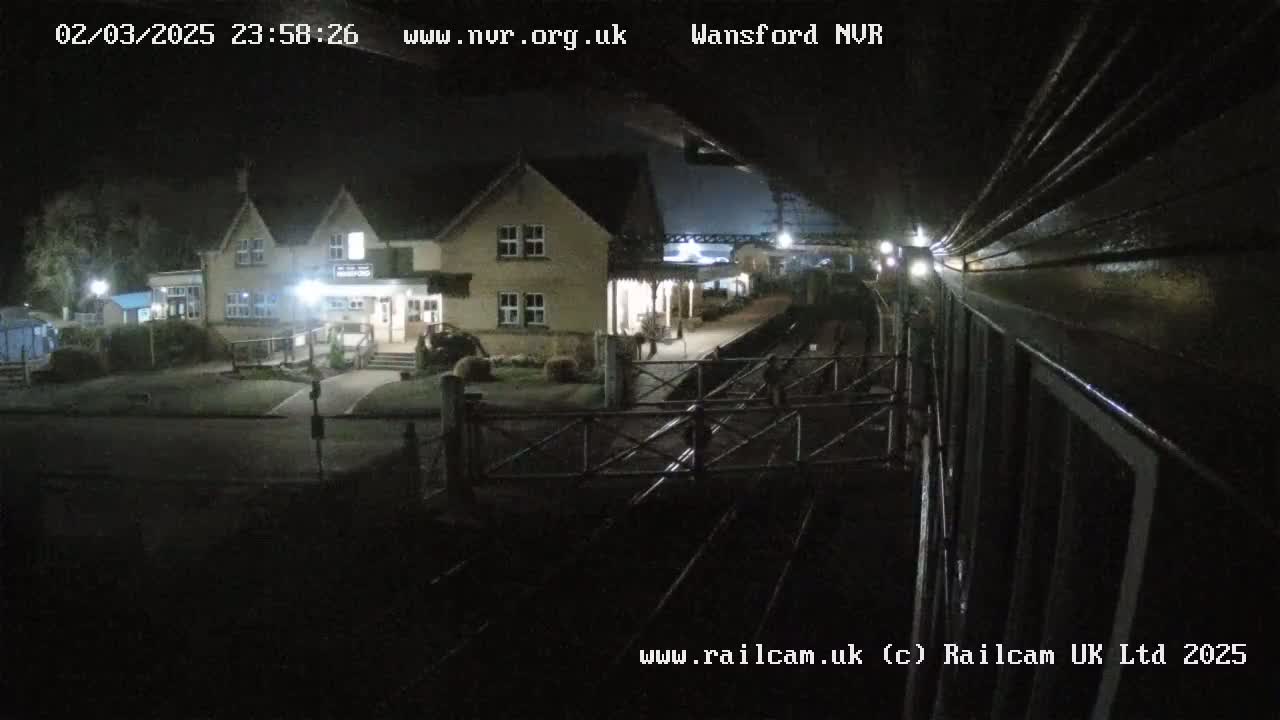 A nighttime view from a train shows a railway station with a building, a platform, and tracks.