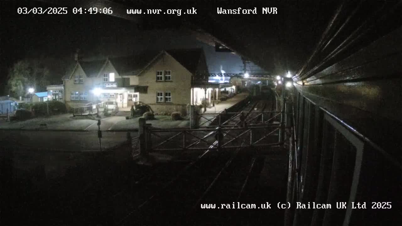 A nighttime view from a train shows a small, illuminated train station building and adjacent parking lot, with train tracks and crossing gates in the foreground.