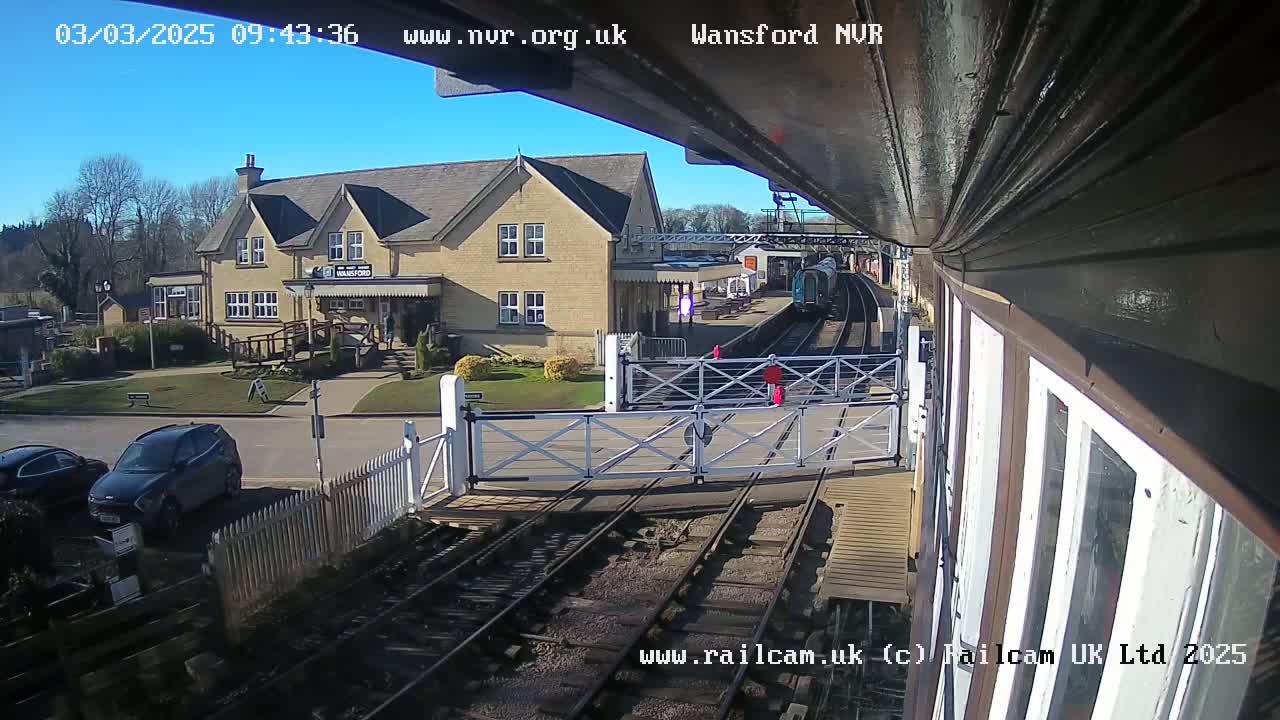 A sunny day at a train station shows a train arriving at the platform, a station building, and several cars parked nearby.
