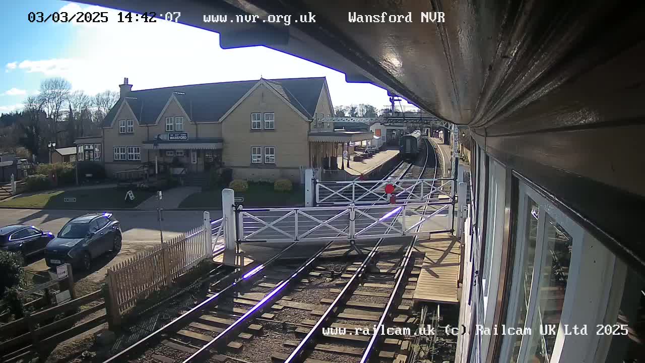 A train approaches a railway station on a sunny day, passing a level crossing with closed gates; a building is visible in the background.