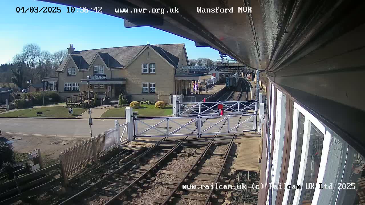 A train is arriving at a station on a sunny day, passing through a level crossing gate near a building.