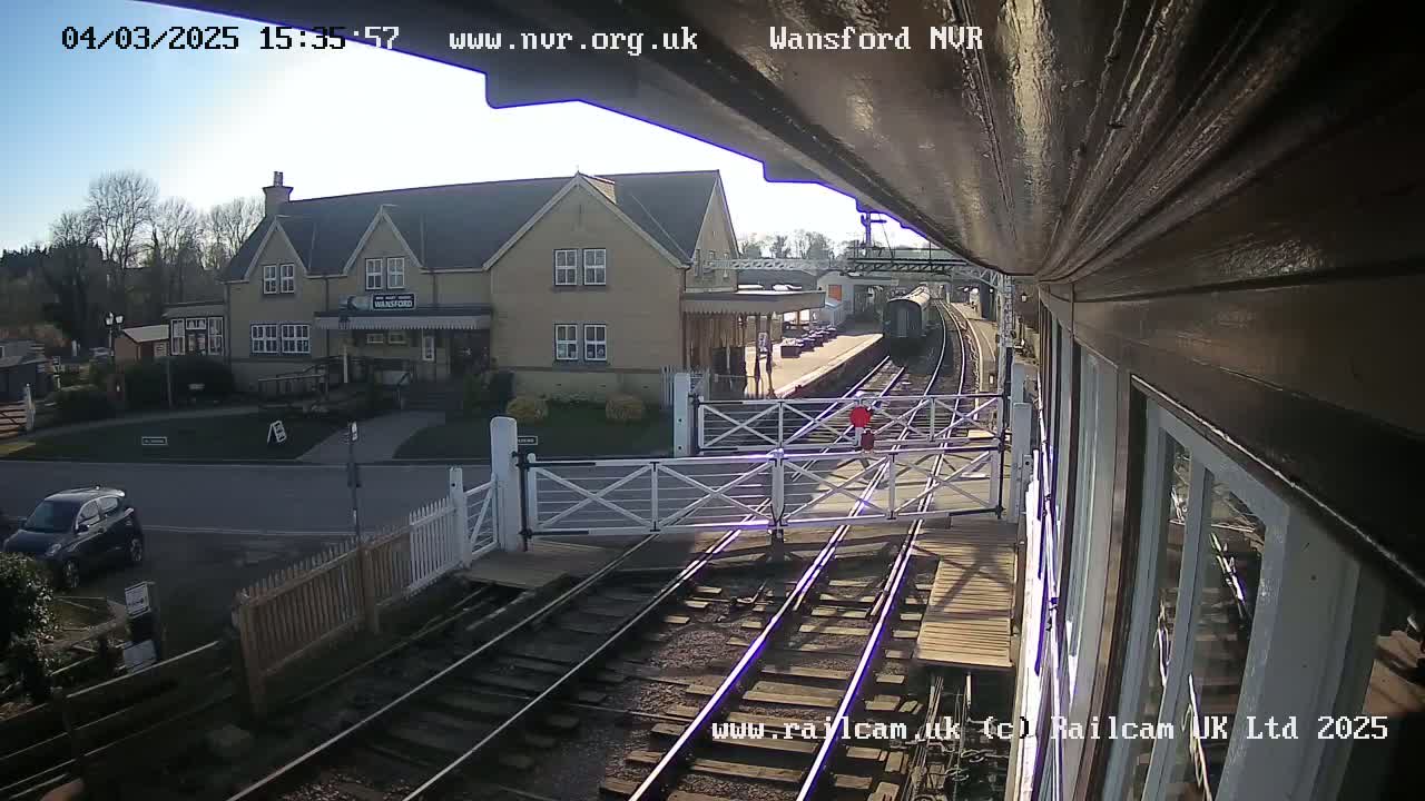A train is approaching a station on a sunny day, passing a level crossing in front of a two-story building.