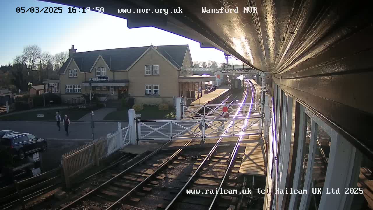 A sunny view from a train platform shows a railway crossing with a train arriving at a station beside a building.