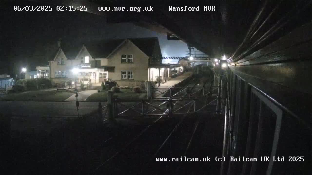 A nighttime view from a train shows a station building with lights on, a gated crossing, and train tracks.