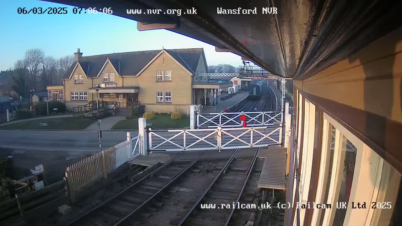 A train approaches a railway crossing in front of a light-colored building under a clear morning sky.