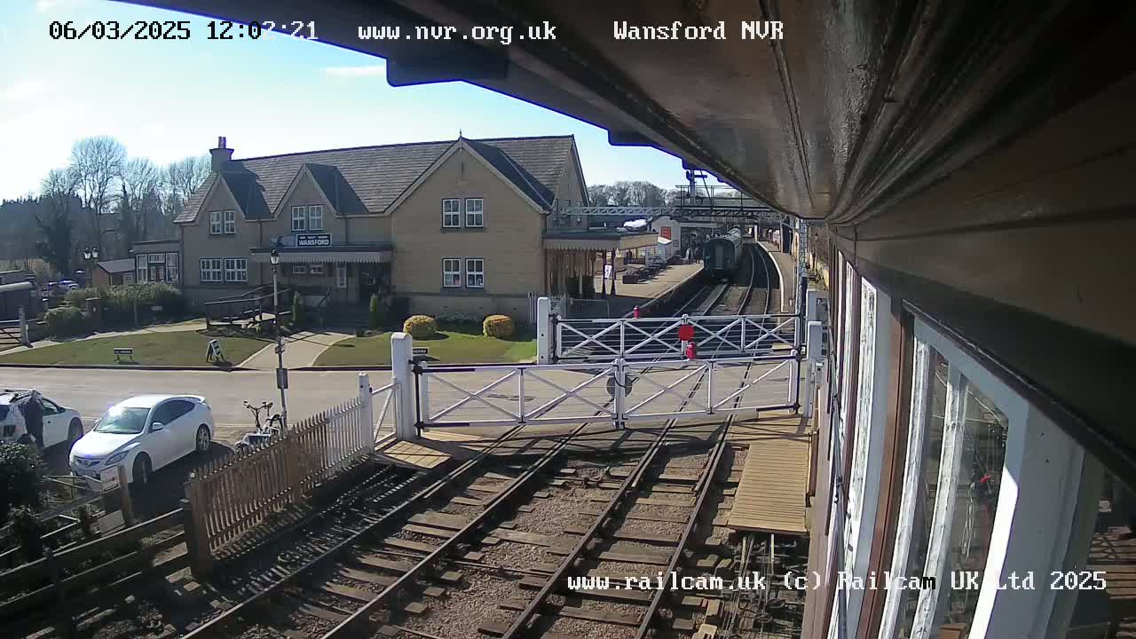 A sunny view from a train platform shows a station building, cars parked nearby, a train on the tracks, and a level crossing gate.
