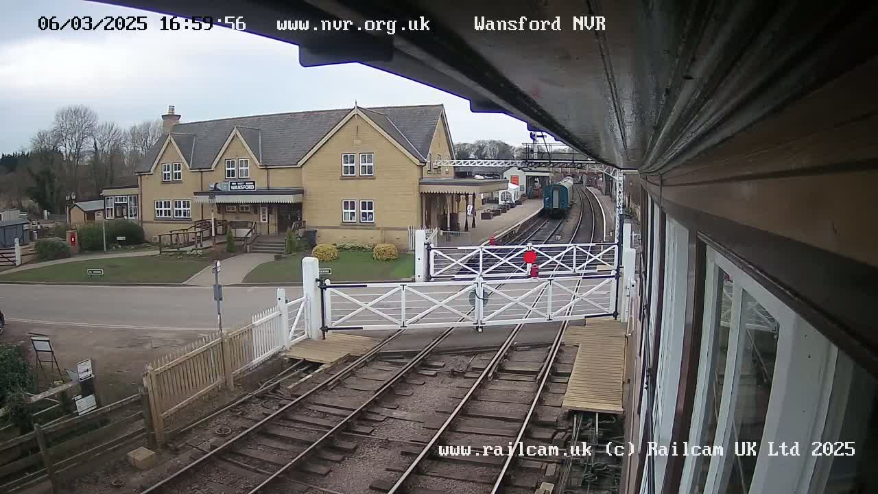 A train approaches a station on a cloudy day, passing through a level crossing gate in front of a building.