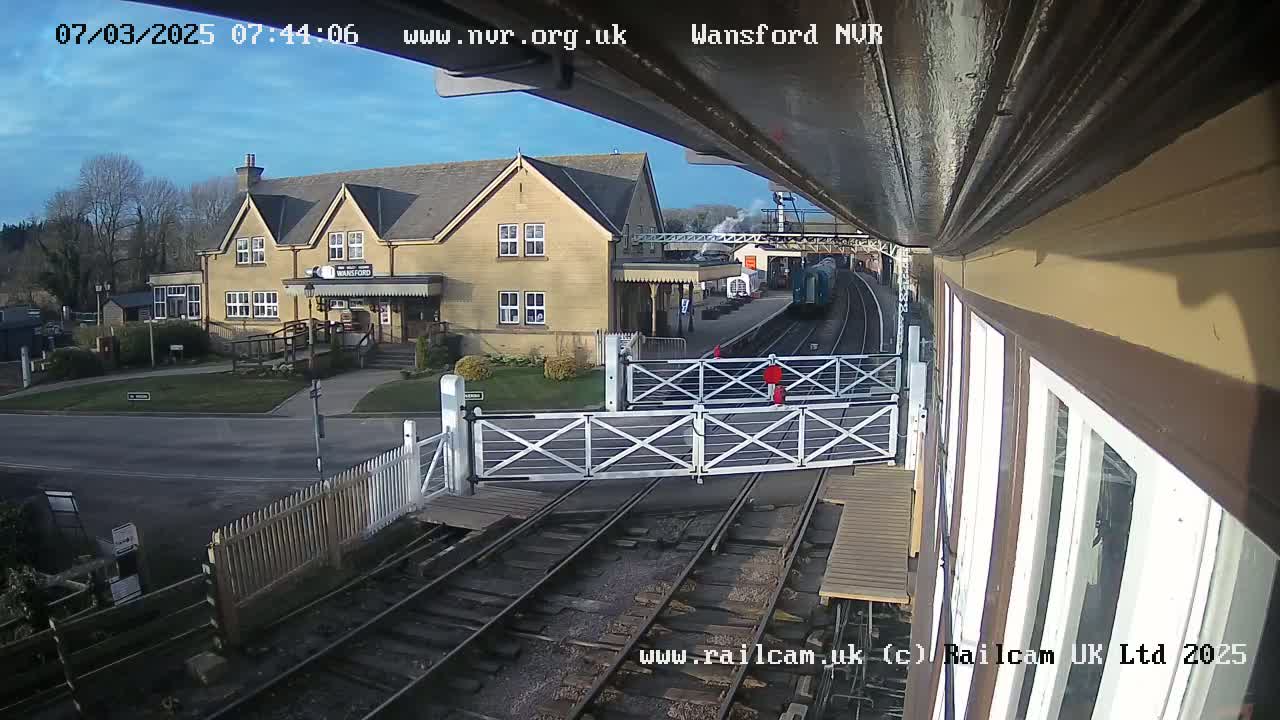 A train is at a station on a sunny day; a building is visible beyond a level crossing.