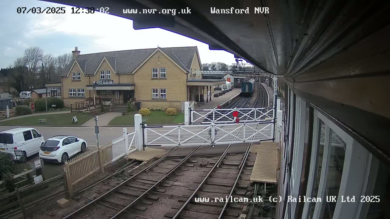 A train pulls into a station on an overcast day, passing through a level crossing with a white gate and two vehicles parked nearby.