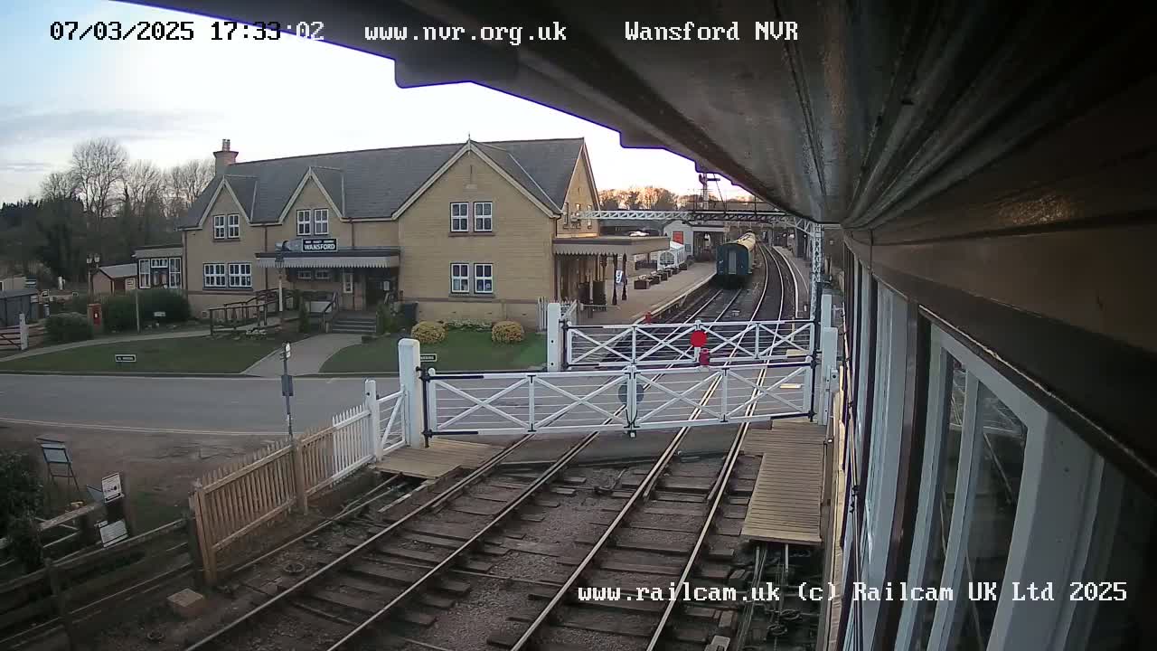 A train is arriving at a station on an overcast day, passing through a level crossing in front of a light-colored building.