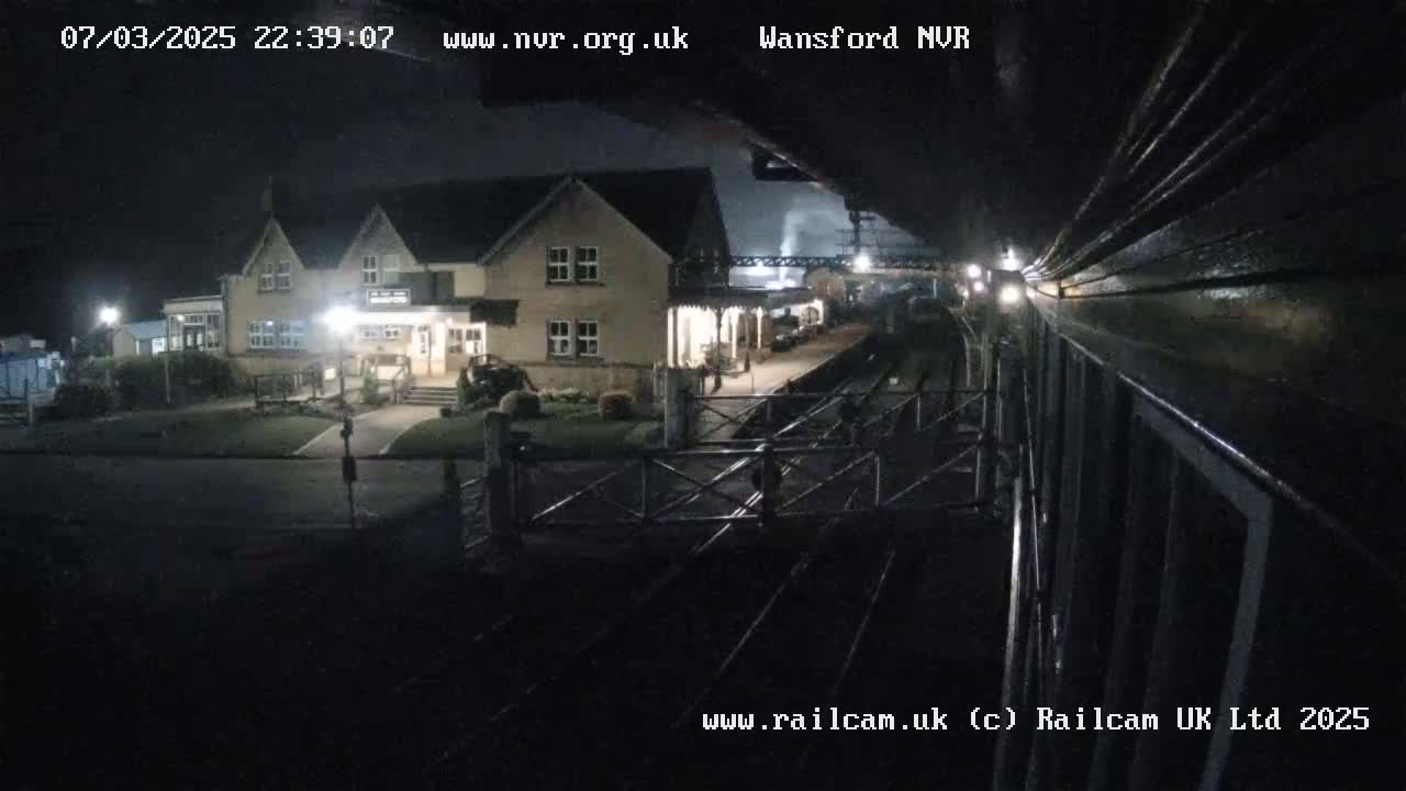 A nighttime view of a train station with a two-story building and train tracks, under dark skies.