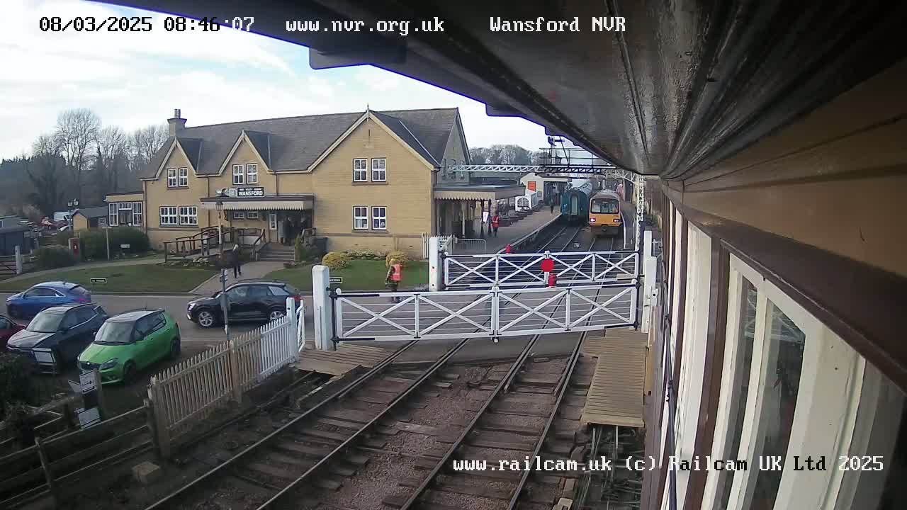 A train passes a railway station on a partly cloudy day, with several cars parked nearby.
