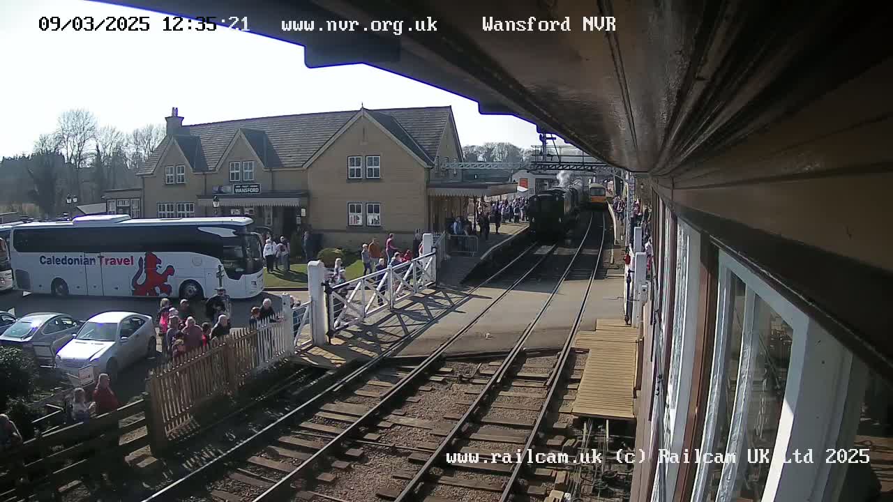 A steam train departs a station on a sunny day, with a crowd of people gathered on the platform and a tour bus parked nearby.