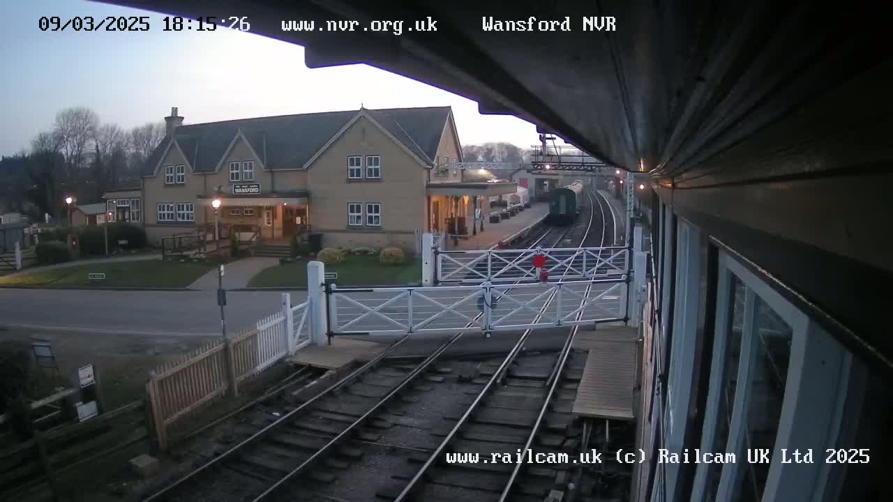 A train approaches a station on a twilight evening, passing through a level crossing gate in front of a building.