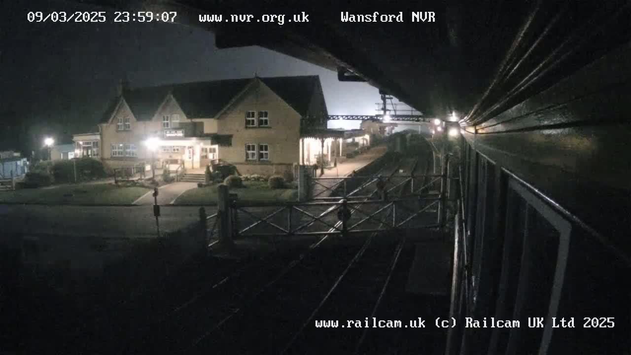 A nighttime view from a train shows a two-story building and a train station with a level crossing gate in the foreground.