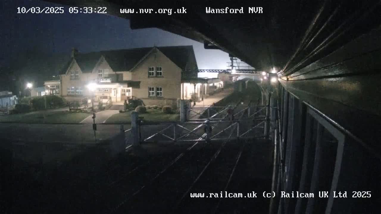 A nighttime view of a train station building and railway tracks, under a dark, possibly cloudy sky.