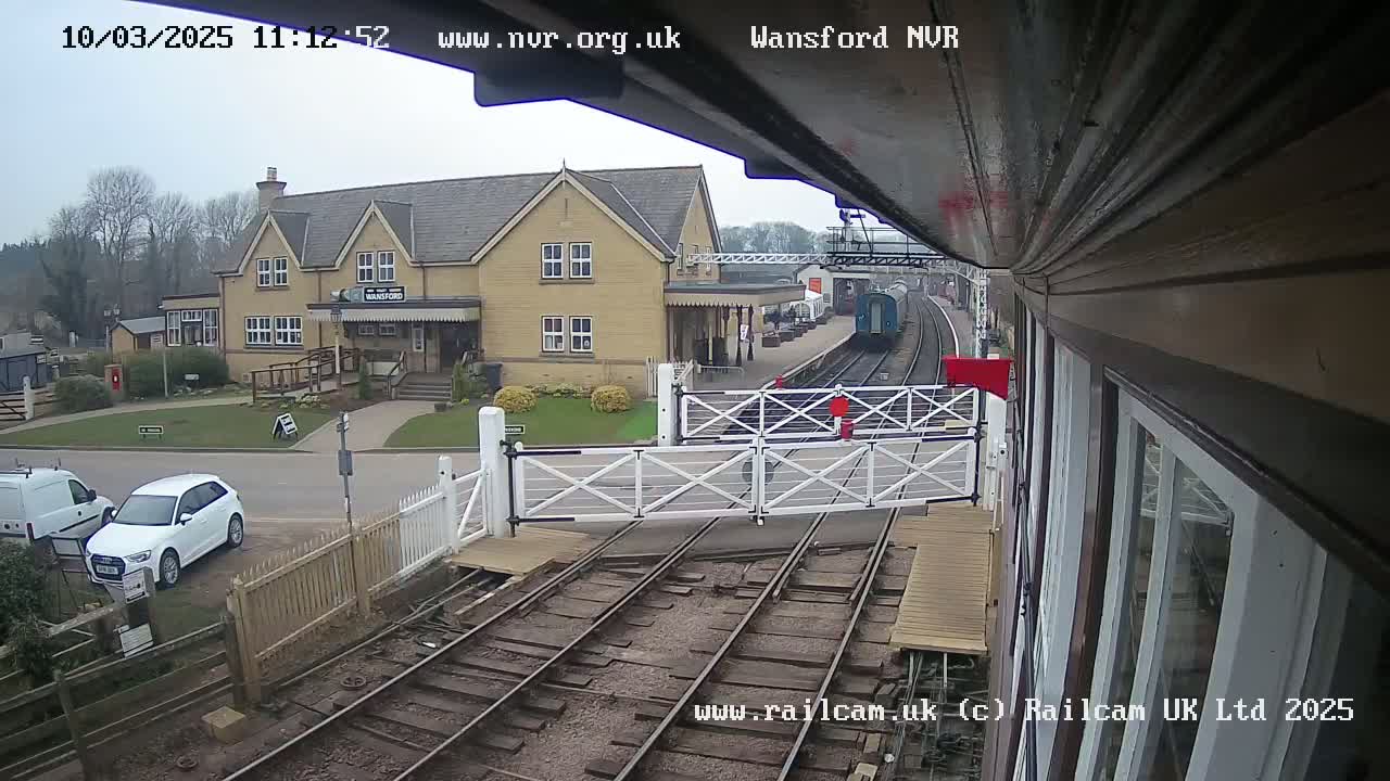 A train arrives at a station on an overcast day, passing through a level crossing with a white gate.