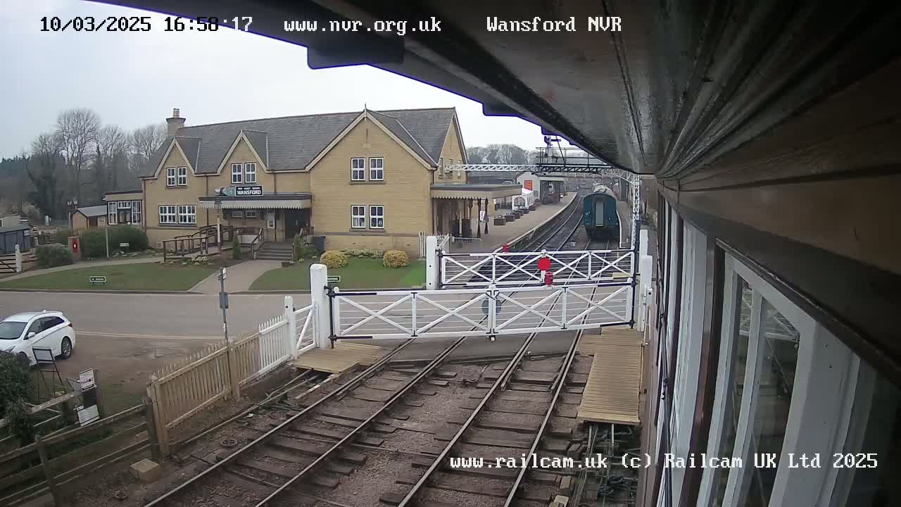 A train is approaching a station on an overcast day, with a white car parked nearby and a building visible in the background.